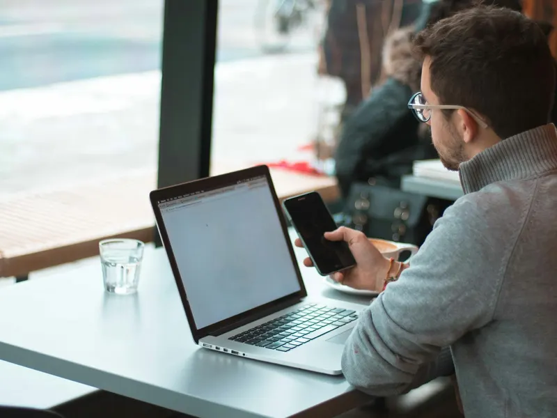 Une personne travaille sur un ordinateur portable à une table près d'une fenêtre, son téléphone posé à côté